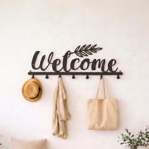 May include: Black metal wall-mounted coat rack with the word "Welcome" in cursive script and a decorative leaf design. A straw hat, beige scarf, and canvas tote bag hang from the hooks. The rack is mounted on a white wall.