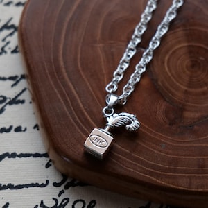 May include: A silver-toned necklace with a pendant shaped like an ink bottle. The pendant has the word "INK" engraved on it. The necklace chain is a twisted rope design, and the background is a wooden surface.