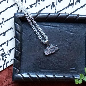 May include: A silver-colored necklace with a pendant shaped like a building, resting on a dark, square tray with a textured surface. The tray is surrounded by a decorative border. The background includes a patterned fabric and greenery.