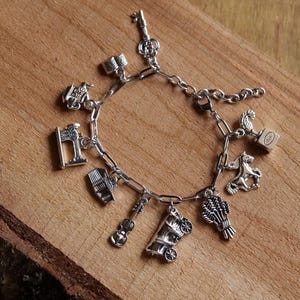 May include: Silver charm bracelet featuring various charms, including a key, a flag, a barrel, a guitar, a covered wagon, a wheat bundle, a horse, and a trophy. The bracelet is displayed on a wooden surface.