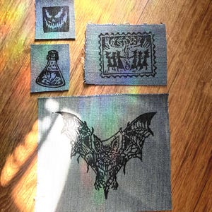 Peut inclure: Quatre écussons rectangulaires en tissu denim avec des motifs imprimés à l'encre noire. Les écussons présentent un visage souriant, une fiole, un groupe de personnages dansant dans un cadre carré et une grande chauve-souris détaillée.