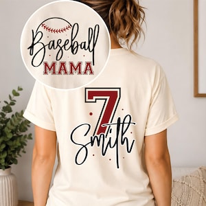 K&ouml;nnte beinhalten: Ein cremefarbenes Kurzarm-T-Shirt mit einem R&uuml;ckendruck, der die Zahl 7 in Dunkelrot mit schwarzer Umrandung, den Namen Smith in schwarzer Schreibschrift und kleine rote Punkte zeigt. Auf der Brust steht Baseball MAMA mit einem Baseball.