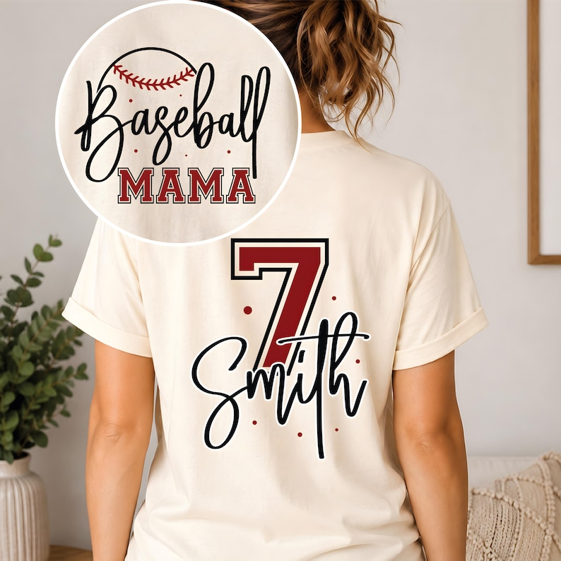 K&ouml;nnte beinhalten: Ein cremefarbenes Kurzarm-T-Shirt mit einem R&uuml;ckendruck, der die Zahl 7 in Dunkelrot mit schwarzer Umrandung, den Namen Smith in schwarzer Schreibschrift und kleine rote Punkte zeigt. Auf der Brust steht Baseball MAMA mit einem Baseball.