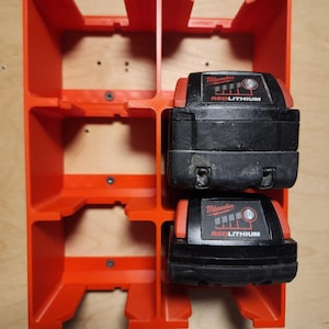 May include: A bright orange plastic storage rack with six rectangular compartments, featuring two black Milwaukee Redlithium power tool batteries inserted into the right-hand side slots. The rack is mounted with screws to a light wood surface.
