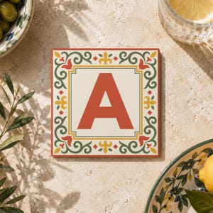 May include: A square coaster featuring a large, bold, terracotta-colored letter A centered on a cream background. The coaster is framed by a decorative border with intricate green scrollwork, yellow fleur-de-lis accents, and terracotta details.