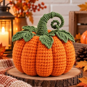 Puede incluir: Una calabaza tejida a ganchillo en un tono naranja vibrante, con una superficie texturizada y acanalada. La parte superior está adornada con varias hojas verdes tejidas y un tallo verde en espiral que se curva hacia arriba.