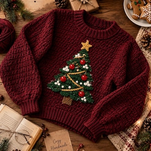 Pode incluir: Um suéter de manga comprida em bordô profundo com um padrão de crochê texturizado e uma aplicação proeminente de árvore de Natal verde na frente. A árvore é decorada com pequenas bolinhas brancas, ornamentos circulares vermelhos e uma estrela.