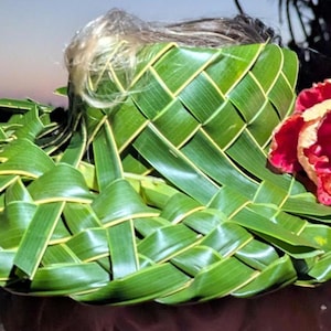 May include: A woven hat made of green leaves, featuring a red and yellow hibiscus flower. The hat's brim is adorned with overlapping leaf sections, creating a textured appearance. The hat is a natural, handmade accessory.