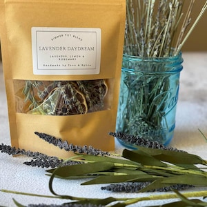 Könnte beinhalten: Ein Kraftpapierbeutel mit der Aufschrift "Lavender Daydream" mit getrocknetem Lavendel, Zitrone und Rosmarin. Ein blaues Einmachglas enthält getrocknete Lavendelstiele. Lavendelzweige und -blätter sind im Vordergrund angeordnet. Der Text lautet "Handmade by Iron & Spice."