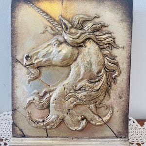 May include: A rectangular, gold-toned relief sculpture of a unicorn's head and flowing mane. The artwork has a textured, aged appearance with cracks and a decorative base. The unicorn's horn is prominent, and the overall style is vintage.