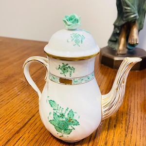 May include: White porcelain teapot with a lid and a green rose-shaped knob. The teapot features green floral designs and gold accents on the handle and spout. The teapot is on a wooden surface.