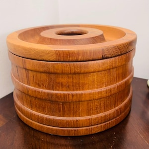 May include: A round wooden container with a lid. The container is made of light brown wood and has horizontal ridges around its body. The lid has a circular indentation in the centre. The container sits on a dark wooden surface.