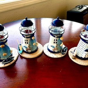 May include: Four decorative lighthouse candle holders. Each lighthouse is white and blue with nautical accents, including small boats and seashell details. The candle holders sit on round, woven coasters and have a dark blue top.