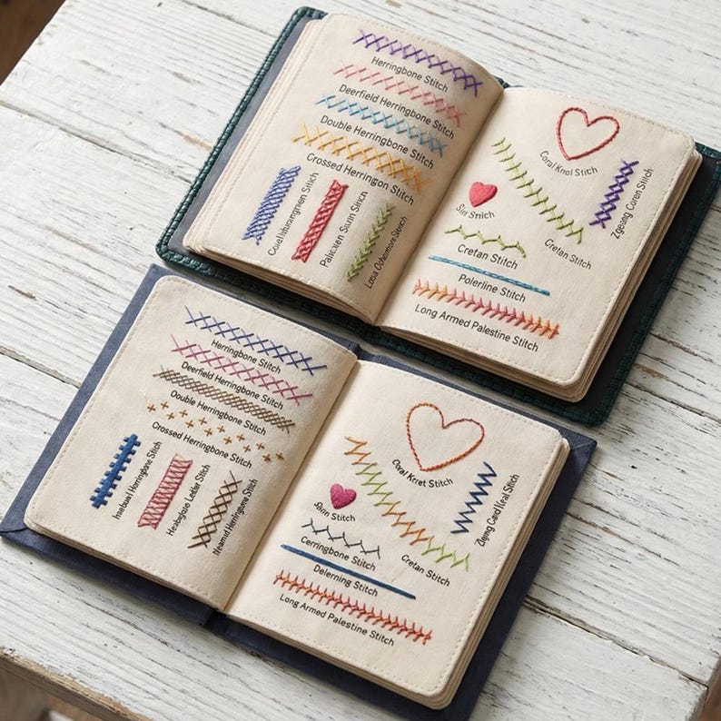 以下が含まれることがあります： さまざまな刺繍ステッチを示す2冊の開いたノート。各ステッチは「Herringbone Stitch」や「Cretan Stitch」などの名前でラベル付けされています。ステッチは、クリーム色の生地に、青、赤、緑など、さまざまな色で施されています。