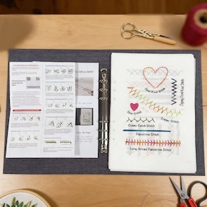 以下が含まれることがあります： 刺繍の指示が書かれた、開いたグレーのバインダー。右側には、ハートやジグザグ模様など、ステッチの例が表示されています。金色のハサミと赤い糸も確認できます。