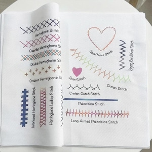 以下が含まれることがあります： さまざまな刺繍ステッチを紹介する白い布サンプル。ヘリンボーンステッチ、クレタンステッチ、コーラルノットステッチなど。青、ピンク、緑、オレンジの色合いのカラフルな糸を使用し、ステッチ名が記載されています。