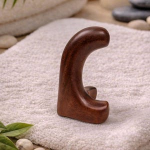 May include: A dark brown wooden massage tool with a curved handle and a base, resting on a white towel. The tool is designed for therapeutic massage. The background includes a lit candle and spa-like elements.