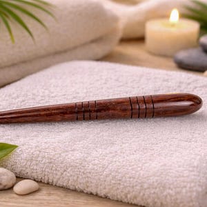 May include: A dark brown wooden massage tool with carved horizontal lines, resting on a white towel. The tool is tapered at one end and rounded at the other. The background includes a lit candle, stones, and a folded towel.