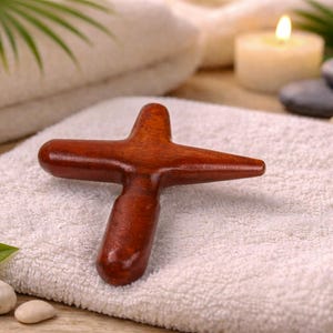 May include: A wooden massage tool in a cross shape, resting on a white towel. The tool is a rich, reddish-brown color. The background includes a lit candle, smooth stones, and green foliage, suggesting a spa or wellness theme.