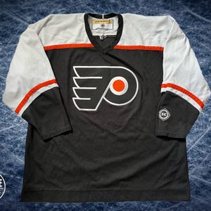 May include: Black and white hockey jersey with orange accents. The jersey features the Philadelphia Flyers logo on the front and a small logo on the sleeve. The sleeves are white with black cuffs and orange stripes. The jersey has a V-neck.