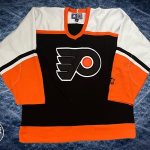 Op de afbeelding: Een ijshockeytrui van de Philadelphia Flyers met een zwarte romp, witte schouders en oranje mouwen met zwarte en witte strepen. De voorkant toont het zwart-oranje gevleugelde logo van het team. Een Starter-logo bevindt zich op de linkermouw.