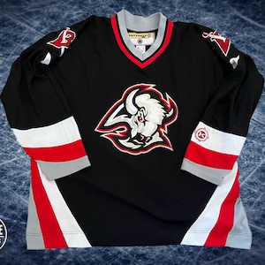 May include: Black hockey jersey with the Buffalo Sabres logo on the front. The jersey has red, white, and gray accents on the sleeves and collar. The sleeves have the letters "B" and "S" on the shoulders. The jersey is made of a breathable material.