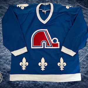 May include: A blue hockey jersey with a white V-neck collar and white cuffs. The jersey features a large red and white logo on the chest, with three white fleur-de-lis symbols at the bottom. The jersey is long-sleeved.