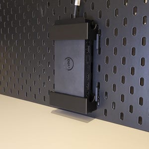 Op de afbeelding: Een zwart Dell-dockingstation bevestigd in een zwarte kunststof houder op een zwart geperforeerd bord. Het rechthoekige apparaat heeft het Dell-logo op de voorzijde en meerdere poorten aan de rechterkant, met een zwarte kabel die aan de bovenkant uitsteekt.