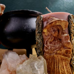 May include: A carved candle featuring a witch with an owl, wearing a hat and dress. The candle is a light brown color with gold accents. A black cauldron and crystals are in the background.