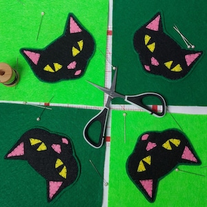 May include: Four black cat head felt cutouts with pink ears and yellow eyes are arranged on green felt squares. A pair of scissors, pins, and a spool of thread are also present, suggesting a crafting project.
