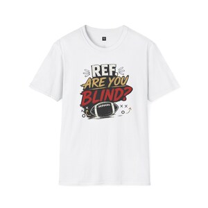 May include: A white short-sleeved t-shirt featuring a graphic print on the chest. The design includes the text REF ARE YOU BLIND? in black and red lettering, accompanied by an illustration of an American football and small decorative markings.