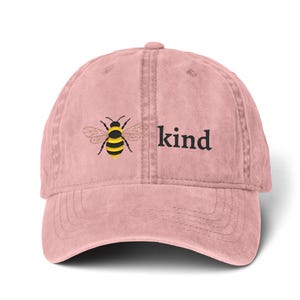 Op de afbeelding: Een roze baseballpet met een gebogen klep. De pet heeft een geborduurde bij en het woord "kind" in het zwart. De bij heeft gele en zwarte strepen. De pet is gemaakt van een zacht, getextureerd materiaal.