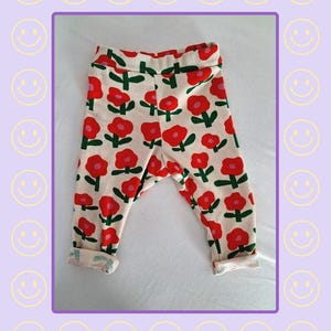 Peut inclure: Pantalon côtelé de couleur crème avec un motif répétitif de fleurs rouges à tiges et feuilles vertes. Le pantalon possède une large ceinture élastique et des ourlets à revers. Le texte MakaStuff est imprimé en violet en bas de l'image.