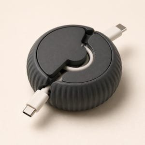 May include: A dark gray circular cable organizer with a ribbed exterior texture. The device features a central rotating mechanism that allows a white USB-C charging cable to be wound inside, with both ends of the cable extending from opposite sides.