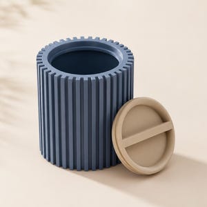 Puede incluir: Un recipiente cilíndrico azul con una textura exterior acanalada verticalmente y una parte superior abierta. Junto al recipiente hay una tapa circular de color beige con una barra de agarre horizontal central.