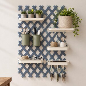 Op de afbeelding: Een blauwe wandorganizer met latwerk, voorzien van meerdere witte planken, twee olijfgroene cilindrische houders en vier metalen haken. De planken bevatten kleine potplanten, een gevlochten mand, een witte vaas, touw en een bril.