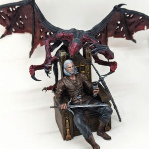 May include: A detailed figurine of a character from The Witcher series, seated on a throne with a sword and goblet. A large, red-winged creature is positioned behind the character. The figure is dressed in brown and black attire.