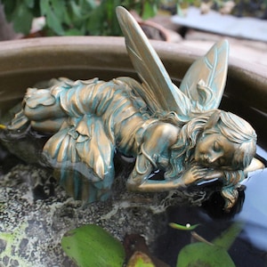 Könnte beinhalten: Bronzefarbene Feenstatue mit türkisfarbenen Akzenten, die friedlich im Wasser ruht. Die Fee hat detaillierte Flügel und liegt entspannt, was ein ruhiges Gartenelement schafft. Ein dekoratives Element für Außenbereiche.
