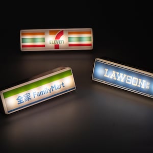 Puede incluir: Tres letreros luminosos con los logotipos de tiendas de conveniencia. El letrero superior muestra el logotipo de 7-Eleven con rayas rojas, naranjas y verdes. El letrero inferior izquierdo muestra el logotipo de FamilyMart con texto verde y azul. El letrero inferior derecho muestra el logotipo de Lawson.