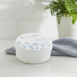May include: A white, round container of Norwex Power Cleaning Paste. The lid features a light blue design with leaves, a sun, and a tree. The container is on a white surface with a gray cloth in the background. The container holds 6.76 fl oz.