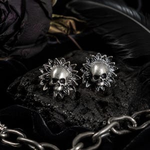 May include: A pair of silver-toned metal stud earrings featuring a central human skull design surrounded by a radiating sunburst or petal-like frame. The earrings have a metallic finish with dark recessed detailing to highlight the skull features.