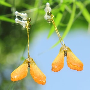 May include: A pair of drop earrings featuring orange and yellow maple seed pods suspended from thin gold-toned metal chains. Each earring includes a small clear plastic post backing and a gold-toned bead at the top of the delicate metal chain.