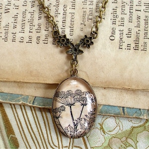 May include: A delicate necklace with a bronze chain and a glass pendant featuring a black and white illustration of a wildflower. The pendant is oval shaped and hangs from a small bronze charm with floral details.