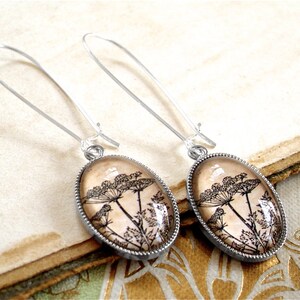 May include: A pair of silver dangle earrings with oval-shaped glass pendants featuring a black and white botanical illustration of Queen Anne's Lace.