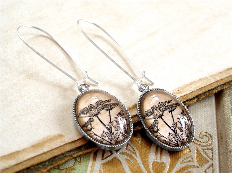 May include: A pair of silver dangle earrings with oval-shaped glass pendants featuring a black and white botanical illustration of Queen Anne's Lace.