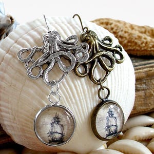Op de afbeelding: Een paar oorbellen met een octopus-thema. Eén oorbel is zilverkleurig, de andere is bronskleurig. Elke oorbel heeft een octopus-bedel en een ronde hanger met een scheepsillustratie. De oorbellen worden op een zeeschelp getoond.