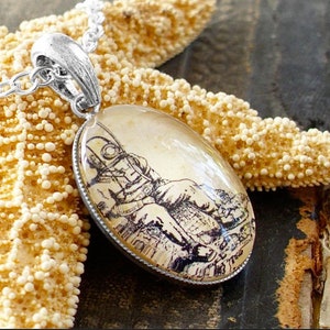 Op de afbeelding: Een zilveren ketting met een ronde hanger met een vintage illustratie van een duiker in een duikpak. De hanger is geplaatst op een beige achtergrond en heeft een zilveren rand.