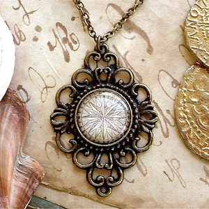 Op de afbeelding: Een gouden ketting met een kompas hanger. Het kompas is omgeven door een decoratief filigraan frame. De ketting ligt op een bruine achtergrond met gouden munten.