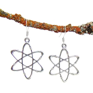 Może przedstawiać: Para srebrnych kolczyków w kształcie atomu wiszących na gałęzi. Kolczyki mają kształt stylizowanego atomu z centralnym okręgiem i krążącymi elektronami.
