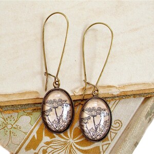 Puede incluir: Un par de pendientes colgantes de bronce con abalorios de vidrio ovalados que presentan una impresión botánica vintage de delicados tallos de flores.